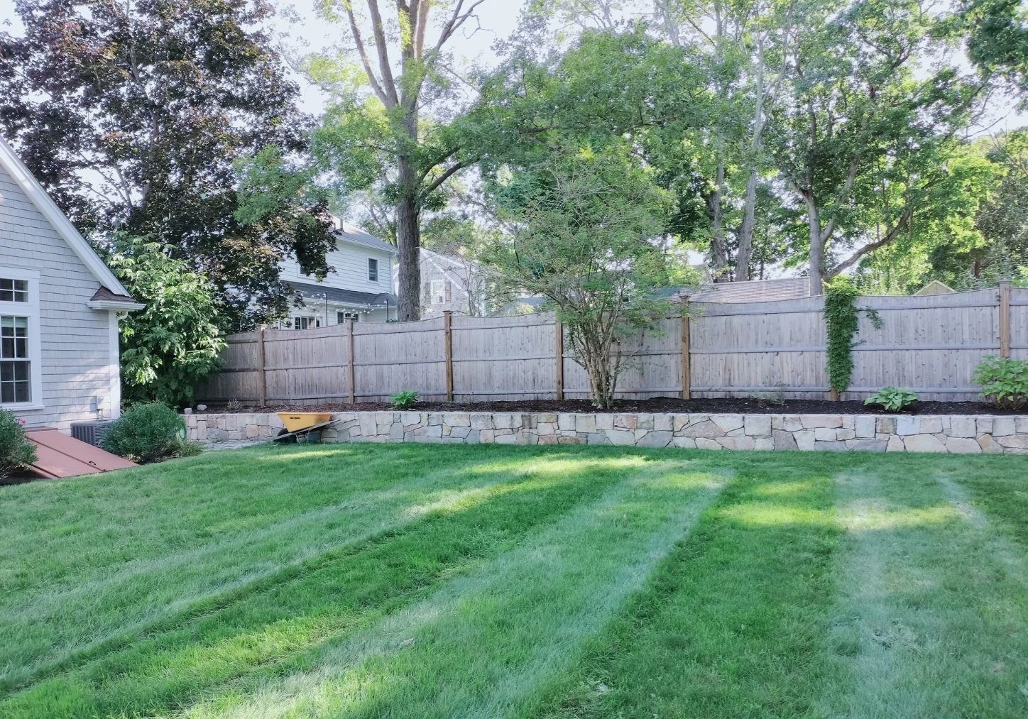 A lawn mowed in stripes below a stone wall planting bed and a wood fence