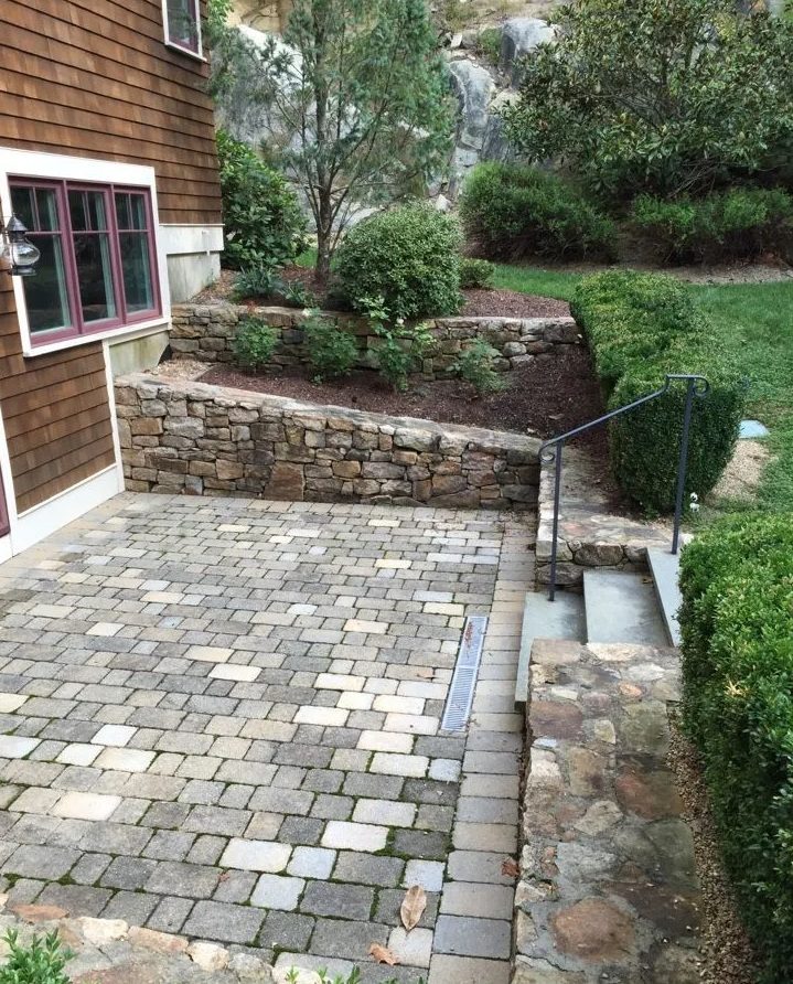 A paver patio with stone steps, a handrail and stone retaining walls beside a house