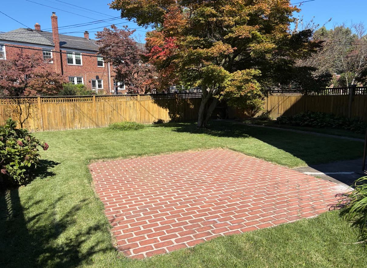 A red brick patio beside a freshly edged green lawn, with a mature tree and fence beyond